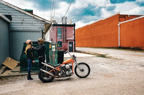 15 Two motorcyclists adjust gear beside an orange vintage motorcycle in an industrial alley.