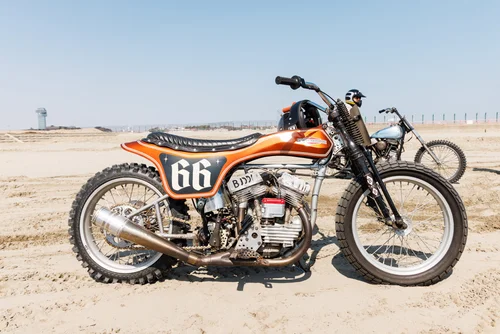1P8A3262 Orange vintage racing motorcycle with number 66 parked on a sandy beach, with a clear blue sky in the background.