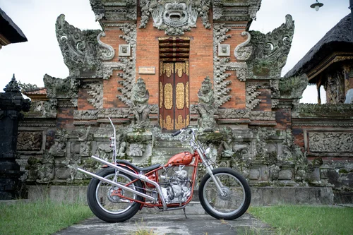 AA-3 Red vintage motorcycle parked on a stone path in front of an ornate Balinese temple gate.