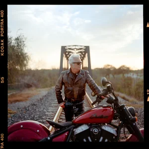 DSC01429-Edit Person in a leather jacket and sunglasses standing beside a red motorcycle on a railroad track, with a metal arch bridge in the background at sunset.