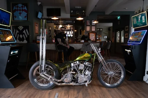 DSCF0402 Custom green motorcycle on a wooden floor inside a dim pub with a bar and patrons in the background.