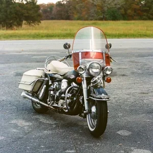 White touring motorcycle with an orange-tinted windshield and saddlebags parked on an empty lot, autumn trees in the background.