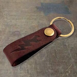 Brown leather key fob with a brass snap and a circular brass key ring on a gray surface, with the word 'BROWN' printed at the bottom.
