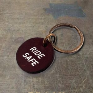 Round black keychain tag with white 'RIDE SAFE' text attached to a metal ring on a gray surface; beige 'BLACK' text appears at the bottom of the image.