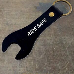 Black bottle opener with a gold ring, stamped with the message 'RIDE SAFE' on a gray surface.
